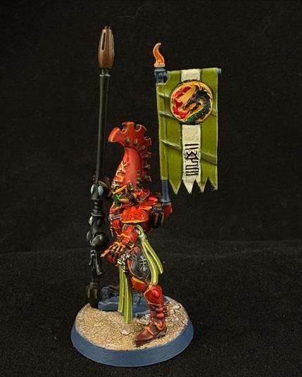 A Warhammer 40K Fire Dragon Exarch miniature. One hand props up a long gun while the other points into the foreground. He wears a back banner which is green with a cream stripe. The banner features runes and a stylized dragon icon.