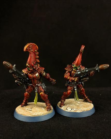 A close up of two Warhammer 40K fire dragon miniatures. They haven’t deep red armor with a pale green tassel hanging from their waist. The one on the right holds his weapon in a ready position while the one on the left has his gun propped against his hip while he prepares to throw a grenade.