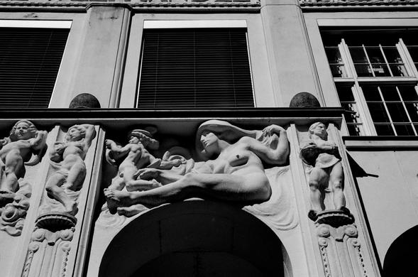 The black and white image shows an ornate architectural facade adorned with classical-style relief sculptures of human figures. At the center, a reclining nude woman with flowing hair is depicted in a graceful pose, flanked by smaller male figures in dynamic stances. The carvings appear to be integrated into the structure above an arched entrance, combining artistic expression with architectural function.