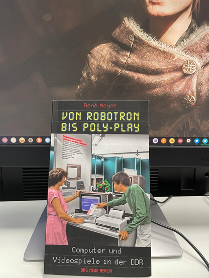Das Bild zeigt ein Buch mit dem Titel "VON ROBOTRON BIS POLY-PLAY" von René Meyer. Das Buch behandelt die Geschichte von Computern und Videospielen in der DDR und trägt den Untertitel "Computer und Videospiele in der DDR - DAS NEUE BERLIN". Auf dem Buchcover sind zwei Personen zu sehen, die an einem Computer arbeiten. Eine Frau in einem rosa Kleid und ein Mann in einem grünen Pullover sind vor einem Computer mit einem Drucker und einem Bildschirm zu sehen. Im Hintergrund ist ein Computerbildschirm mit einem Spiel zu sehen. Das Buch liegt auf einem Tisch vor einem Computermonitor, der ebenfalls ein Spielbildschirm zeigt.