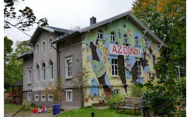 Dreistöckiges Haus, mit bunter Fassade auf der steht "AZ Conni".
Eine Sitzbank vorne rechts, dazu Bamue an deren Seiten