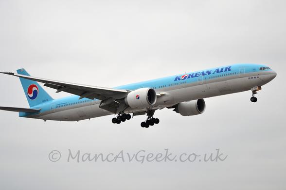 Side view of a light blue, twin engined jet airliner flying from left to right at a low altitude, with undercarriage lowered and flaps extended from the rear of the wings, suggesting it is about to land.
Th top half of the plane is light blue, with a thick grey stripe separating that from the white belly.
There are dark blue "Korean Air" titles on the upper forward fuselage, a slightly modified, red, blue, and white Yin-Yang symbol replacing the letter "O".
That same symbol appears on the tail, and on the sides of the white engine pods under the wings.
Flat grey sky fills the background.
