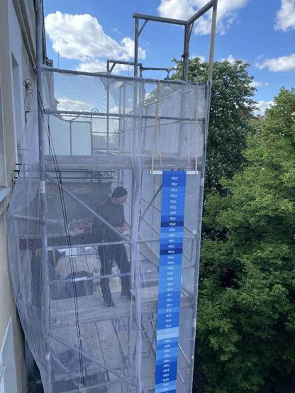 ein in weißer Gaze gehülltes schmales Baugerüst vor dem obersten Geschoß eines Gründerzeitwohnhauses. Davor hängt ein schmales Banner nit den sog. "Klimastreifen". Auf einer Plattform ist eine männlich gelesene Person beim Aufbau beschäftigt. Blauer schönwetterbewölkter Himmel