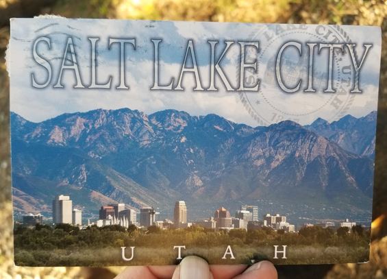 A postcard from Salt Lake City. The image shows mountains - part of the Rockies. In the foreground, with the mountains as a backdrop, is the city skyline. Text in the sky reads Salt Lake City and text at the very bottom says Utah.