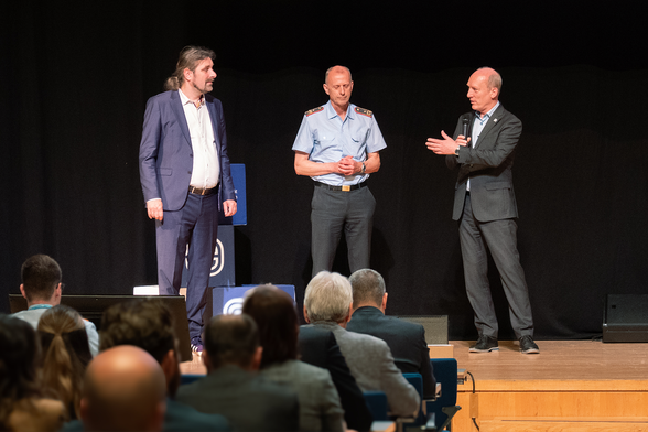 Das Bild zeigt die Eröffnung des Symposiums SIC!2025 in der Händelhalle Halle (Saale). Im Mittelpunkt der Bühne stehen drei Männer nebeneinander vor einem dunklen Bühnenhintergrund. Links im Bild steht Prof. Dr. Christian Hummert, Forschungsdirektor der Cyberagentur, im blauen Anzug mit offenem Hemdkragen. In der Mitte befindet sich Generalmajor Dipl.-Inf. Jürgen Setzer, Stellvertreter des Inspekteurs Cyber- und Informationsraum der Bundeswehr, in blauer Uniform mit Schulterklappen und verschränkten Händen. Rechts steht Wilfried Karl, Präsident der Zentralen Stelle für Informationstechnik im Sicherheitsbereich (ZITiS), in dunklem Anzug mit Mikrofon, in einer erklärenden Gestik. Im Vordergrund sind Teilnehmende des Symposiums von hinten zu sehen, die aufmerksam auf die Bühne blicken. Die Szene wirkt konzentriert und formell, und dokumentiert den offiziellen Auftakt der Veranstaltung. Foto: Cyberagentur