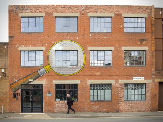 Looking front on at a red brick building. It is an industrial building with three floors. The ground floor has a door on the left hand side and three windows to the right of them. The first and second floors have four windows each. A man is walking past on the pavement. One of the first floor windows has a piece of street art painted around it - a large magnifying glass. The second window from the left is in the lens of the magnifying glass. The handle goes down to the ground floor, over and to the left of the doors.