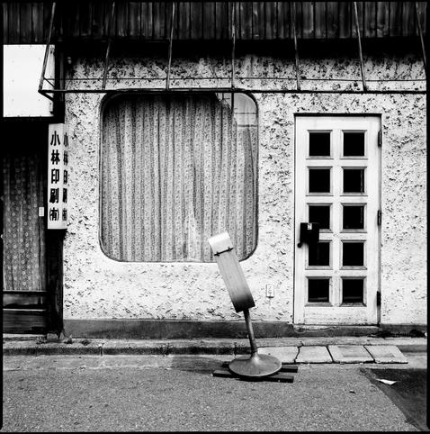 black and white square photograph of a window. the window is large and square with rounded corners. the building is retro looking with fake stucco. a sign to left of the building has japanese characters "小林印刷（有)" (kobayashi printing ltd).