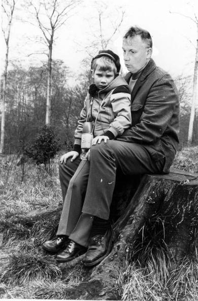 an old black-and-white picture. a middle-aged man sits on a tree stump on a wooded hill. between his legs stands a boy of about four, who looks sadly into the camera. The man speaks to him comfortingly