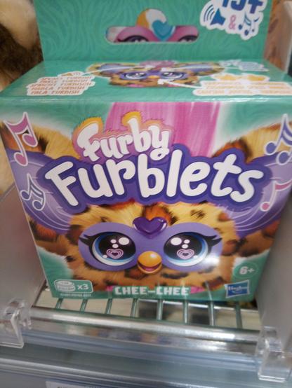 Ein Regal mit einer türkisfarbenen Verpackung und einem kawaiiigen Gesicht eines lütten Furbys aufgedruckt. Dazu steht: Furby Furblets" Und unten – wohl der Name des Furblets: "Chee Chee".