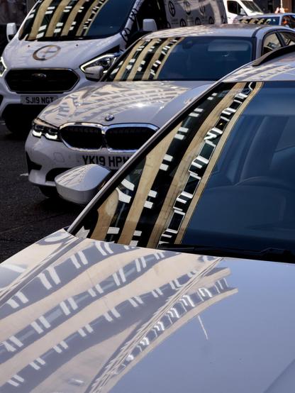 The brightly-lit reflection of a city centre building is caught in the bonnets and windows of two cars and a van in a shady side street