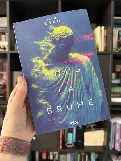 Livre pris en photo devant une bibliothèque. 
La couverture de Sous la brume montre une statue antique dont une partie s’effrite en poussières. Des artéfacts de glyphes apparaissent aussi sur l’image.