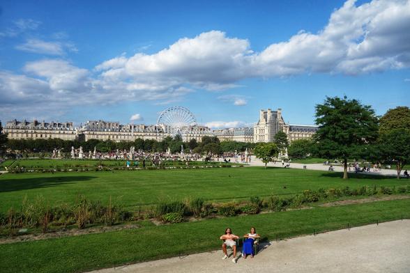 The image shows a wide, green lawn in what appears to be a park, possibly the Tuileries Garden. In the background, beyond the green space and trees, there's a busy crowd of people strolling through the park, with a large white Ferris wheel and a grand, light-coloured building with ornate architectural details further behind. The sky is bright blue with fluffy white clouds. In the foreground, two people are sitting on lawn chairs on a dirt path beside the green lawn. The overall scene captures a vibrant park atmosphere on a sunny day.