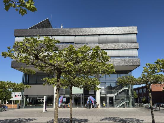 Ein modernes mehrstöckiges Gebäude mit einem gläsernen Eingang, umgeben von grünen Bäumen. Die Fassade weist eine Kombination aus dunklen und hellen Materialien auf, und darüber ist ein blauer Himmel zu sehen. In der Nähe des Eingangs sind Menschen versammelt, und in der Nähe sind Fahrräder geparkt.
Wissenschaftszentrum in Kiel - Veranstaltungsort.