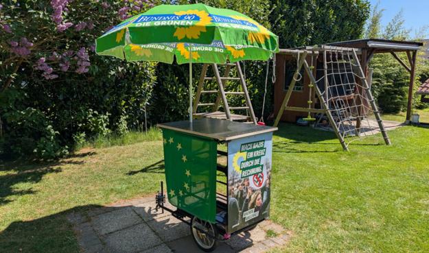 Ein Fahrradanhänger als Wahlinfostand mit einem Stehtisch und integriertem Sonnenschirm inkl. Werbung für die Grünen.