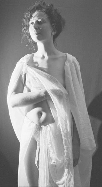 Black and white image of a person in a flowing white garment, striking a contemplative pose with one arm wrapped around their body. The background is neutral, creating a soft focus on the subject's expression and silhouette. Their face glows.
