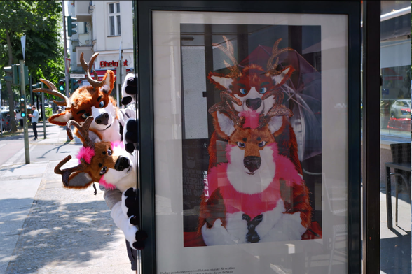 Two Deer-Fursuiters  looking from behind a Bus-Stop. The Bus Stop features an Image with both of them instead of a boring advertisement.