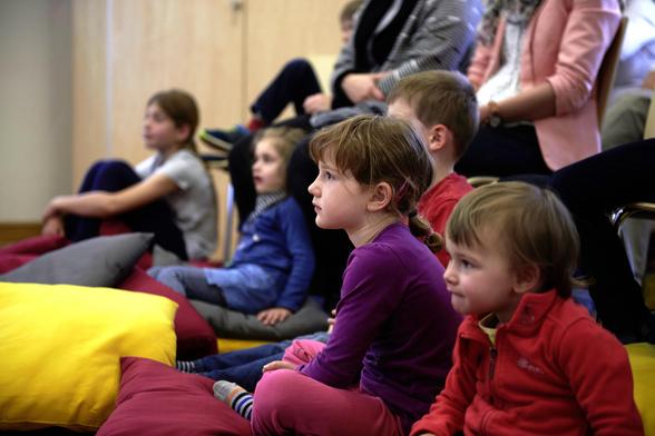 Kinder verschiedenen Alters sitzen für eine Vorlesestunde auf dicken Kissen auf dem Boden und schauen nach vorne. Dahinter sitzen Erwachsene auf Stühlen.