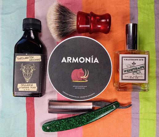 (Le Chat Mistral description)

The image showcases a collection of grooming items arranged on a striped background. At the centre, there's a round tin labelled "Armonía" with an image of a mammoth. To the left, there's a dark bottle labelled "Opulence Aftershave Splash." Above the tin, there's a shaving brush with a red handle. To the right of the tin, there's a small bottle of perfume labelled "Weinstrasse." At the bottom, there's a green glittery straight razor. The items are neatly organised, suggesting a traditional shaving setup.