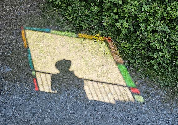 The image of a window and balcony is cast onto the ground by bright sunshine from behind. The window is surrounded by coloured glass and a shadow figure in a hat is standing at the window.