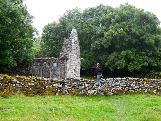 eine wiese … eine person steigt über die treppe in einef natursteinmauer um die ruine einer alten kirche ohne dach anzusehen … im hintergrund große bäume, die zum teilmdie sicht auf die ruine verdecken