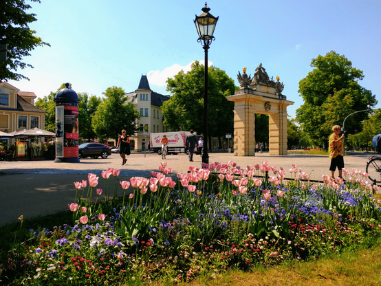 Das Jägertor in Potsdam steht umringt von sorgfältig gepflegten Blumenbeeten, wo unter anderem pinke Tulpen wachsen. Es ist warmer Frühsommer, Menschen schlendern mit Eis in der Hand oder dem Fahrrad dabei über den kleinen Platz. DIe Sonne scheint von oben, es sind wenige Wolken zu sehen, und das Tor, die Blumen und der sommerliche Schattenwurf machen ein kontrastreiches Bild.