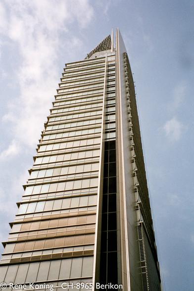 The image features a tall, modern skyscraper reaching towards the sky. The building has a sleek, contemporary design with a predominantly glass facade, reflecting the surrounding environment. The structure is composed of numerous horizontal and vertical lines, creating a rhythmic pattern. The windows are arranged in a grid-like pattern, with some sections featuring darker glass, possibly for shading or aesthetic purposes. The building tapers towards the top, culminating in a pointed architectural feature. The sky in the background is partly cloudy, with a mix of blue and white hues. The image is taken from a low angle, emphasizing the height and grandeur of the skyscraper.