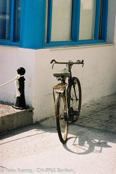 Das Bild zeigt ein altes Fahrrad, das vor einer weißen Wand mit blauen Fensterrahmen geparkt ist. Das Fahrrad hat einen grünen Sattel und ein Nummernschild mit der Aufschrift "-950". Es steht auf einem gepflasterten Bürgersteig und wirft einen Schatten auf den Boden. Links neben dem Fahrrad befindet sich ein schwarzer Poller, um den ein weißes Seil gebunden ist, was auf einen Sperrbezirk hindeutet. Die Wand hinter dem Fahrrad ist weiß mit sichtbaren Gebrauchsspuren, und die Fenster haben blaue Rahmen. Die Szene ist gut beleuchtet, was auf einen sonnigen Tag hindeutet. In der Gesamtkomposition steht das Fahrrad im Mittelpunkt, wobei das Gebäude und der Bürgersteig den Kontext bilden.