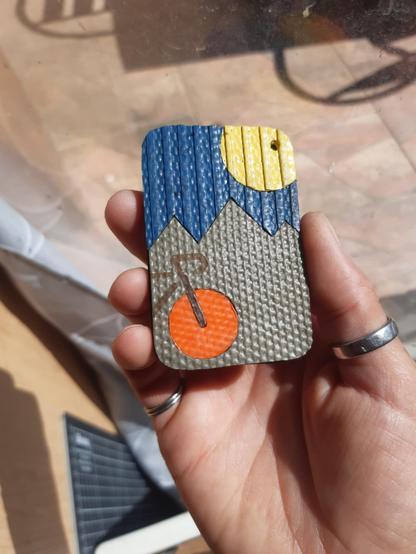 Porte-clés rectangulaire réalisé à partir d'une couche de chambre à air de tracteur prise en sandwich entre 2 couches de tuyaux de lances à incendie coupés pour former :
- des montagnes kakies
- un ciel bleu
- un soleil jaune
et au 1er plan, un vélo gravé au laser, avec une roue en tuyau orange