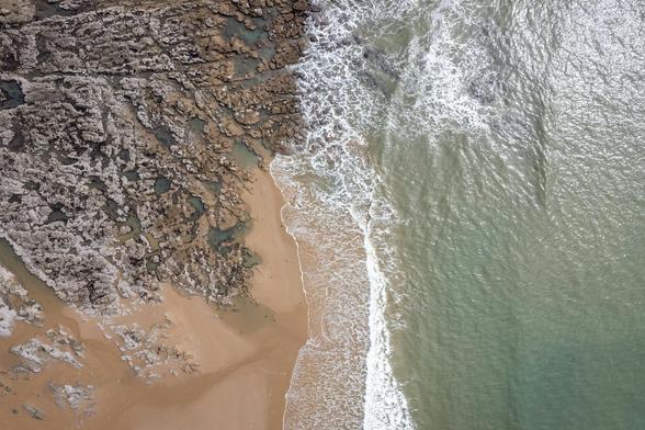 Aerial view of a beach, with sand and rocks on the LHS and sea on the RHS.