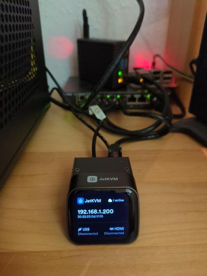 A turned on JetKVM device on top of a wooden desk. On the left there is a part of a black computer case and it's fan grid visible. In the background there is a 8 port ethernet switch, one Raspberry Pi with a case and one Raspberry Pi without a case.

The JetKVM device has a small display showing its IP and MAC address. It can be used to remote control the attached server via USB mouse or keyboard and see its screen via HDMI.