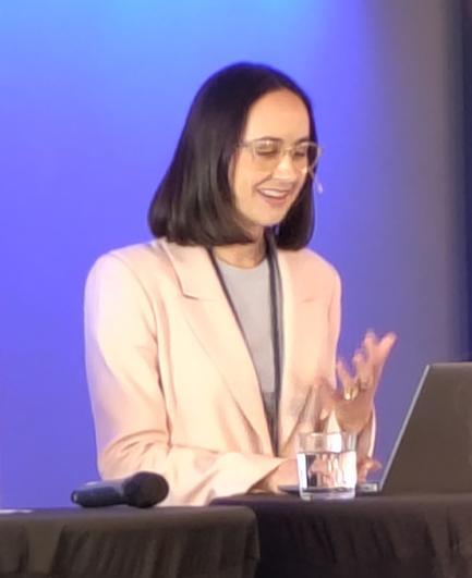 Ashley Cooper at the EASE conference podium. She's a smiling white woman with shoulder length black hair and glasses, wearing an off-white jacket and a light grey top.