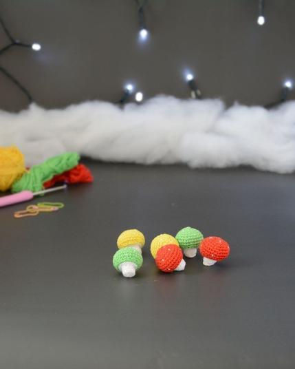 Six petits champignons crochetés en coton coloré. Leurs chapeaux sont rouge, jaune et vert. Ils sont posés sur un fond gris foncé. En arrière-plan, des pelotes et un crochet sont partiellement visibles, ainsi qu'un peu de rembourrage blanc et une guirlande lumineuse.