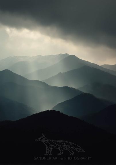 A moody photograph of layered mountain ridges under a stormy sky, with beams of light breaking through the clouds, casting a mystical glow over the dark, forested slopes. A geometric fox logo and the text "Sandner Art & Photography" are overlaid in the bottom right corner.