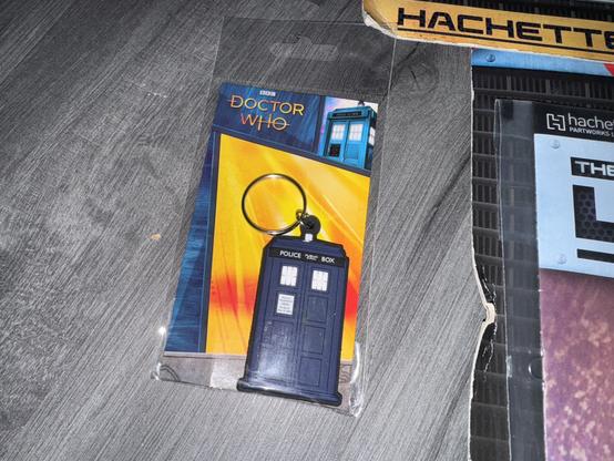 A Doctor Who TARDIS keychain in packaging. The background features a colorful design, highlighting the iconic blue police box. Other items partially visible include Hachette branding.