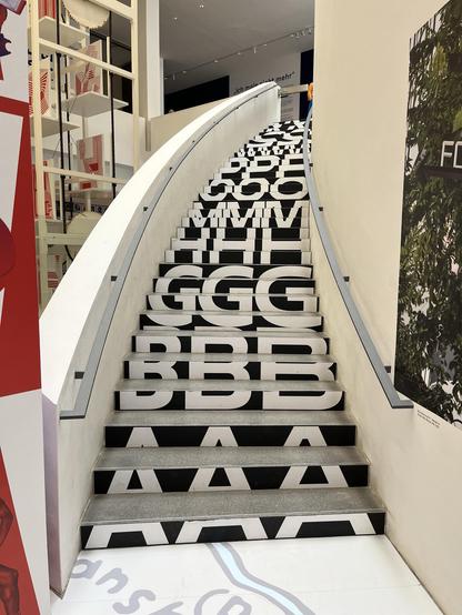 A curved staircase featuring large black and white letters printed on the steps forming the start of the alphabet. The environment appears modern, with additional design elements visible in the background.