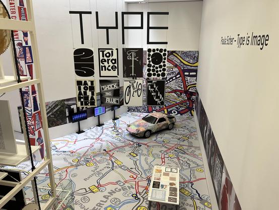 The image depicts an art installation titled "Type is Image" by Paula Scher. It features a vibrant floor design resembling a map, along with various oversized typographic displays hanging from the ceiling.