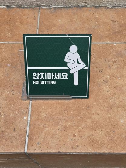 A green sign with an pictogram of someone sitting and English and Korean text saying don't sit