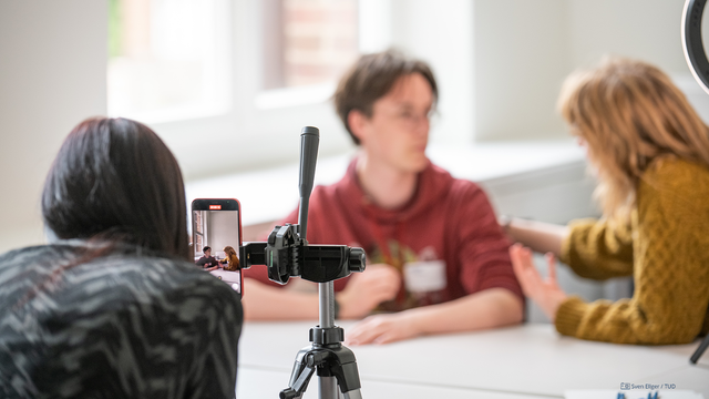 Foto von einer Gruppe aus drei Personen, die mit einem Smartphone ein Video aufzeichnen. Eine Person ist dabei hinter der Kamera. Die beiden Personen, die gefilmt werden, sind unscharf abgebildet.