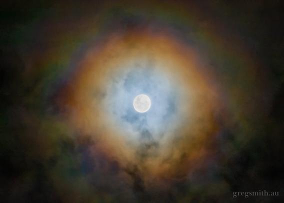 An almost-full moon through clouds, producing an orange glow and a rainbow.