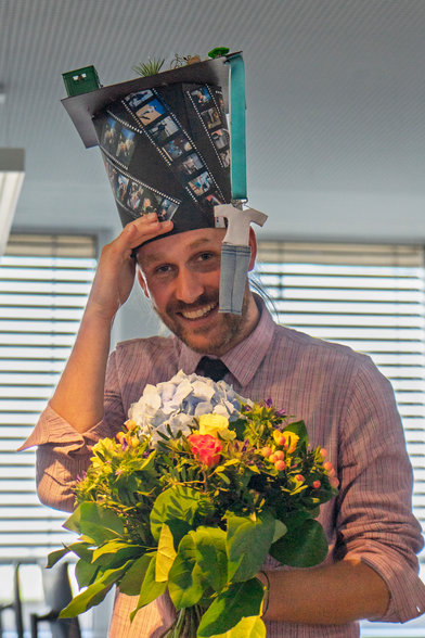 Moritz Epke with a crafted PhD hat with photo strips and a miniature T-Shirt and jeans ribbon hanging from the side of the hat. He is holding a bouquet of orange, yellow and white flowers.