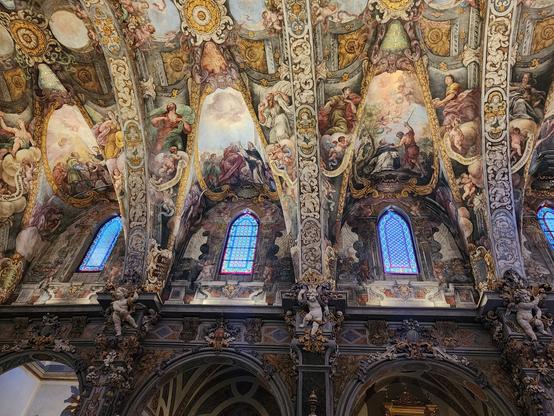 three small, vaulted stained glass windows high above viewer. the windows are of simple floral/abstract designs and the walls are covered with elaborate frescoes of saints and angels.