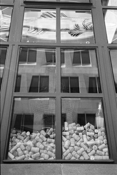 A black and white photograph captures a shop window. The upper section features a stylized drawing of a fish on a plate. Below, the window reflects buildings across the street. The lower portion is filled with numerous wine corks, and at least one empty glass bottle rests among them. The combination of the fish artwork and the wine corks suggests the establishment might be a seafood restaurant or a wine bar. The reflections of the surrounding buildings provide additional context. The photograph is taken from a slightly low angle, directing the viewer's gaze towards the window display. The varying textures, from the smooth glass and detailed drawing to the irregular arrangement of the corks, are emphasized by the monochrome rendering. The presentation appears deliberate, aiming to attract the attention of passersby.