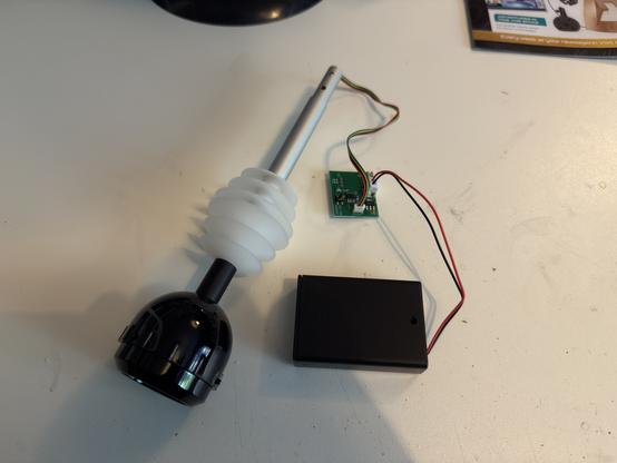 An assembly of electronic components, including a white flexible boot, a black actuator or housing, a printed circuit board (PCB) with wires, and a black battery holder. These items are placed on a white surface.