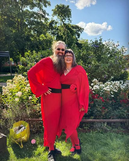 The image features two individuals standing outdoors in a park setting. They are both wearing matching red outfits with long sleeves and black belts. The person on the left has a long beard and is wearing sunglasses, while the person on the right is wearing glasses. The background includes lush greenery and blooming flowers, with a clear blue sky and some clouds visible. The scene is bright and sunny, suggesting a pleasant day.