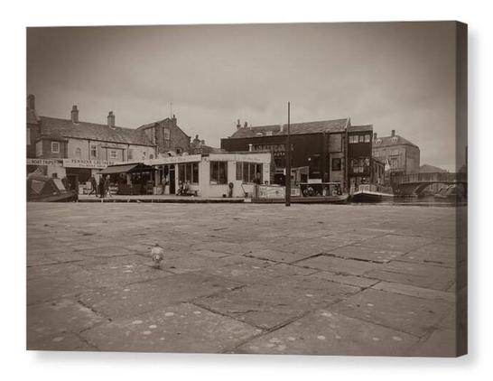 Black and white photograph showing a canal cafe in the background with a flagstoned pavement, and pigeon, in the foreground.  The image is shown printed upon a box canvas.