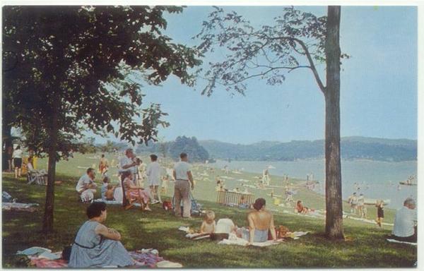 Color image of sunbathers and swimmers on a 1960s beach near a lake. Image shows groups of picnickers, sunbathers, and swimmers on a lawn above a sandy beach leading into the water at Atwood Lake, Carroll County, Ohio, USA, circa 1960s. Trees dot the shoreline and beach area, and people are dress in 1960s era clothing, from sundresses to bikinis and Bermuda shorts, on blankets and beach towels, with the tones of the image reflecting the style of photofinishing from that era.