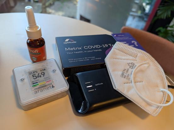 A conference table with a CAN95 ear loop mask, an aptitude metrix reader and COVID test cartridge box, an aranet4 CO2 meter reading 549, and an orange Profi nasal spray bottle.