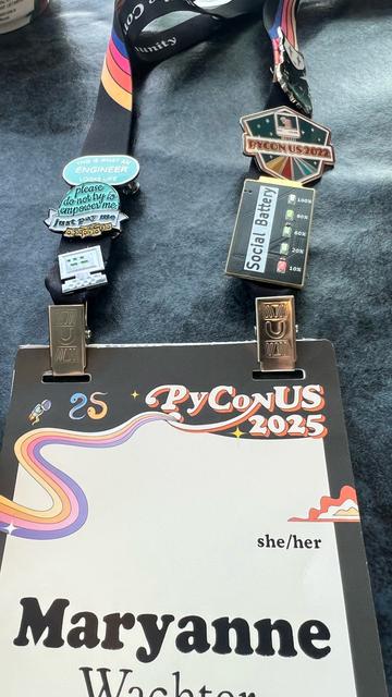 A pyconus conference badge adorned with various pins including one that is a social battery indicator with leds