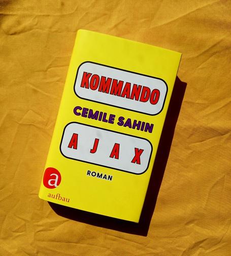 Foto des Buches, auf dessen knallgelbem Cover in zwei weißen, schwarz umrandeten Flächen in roter Schrift der Titel steht, dazwischen in Lila der Name der Autorin. Es sieht ein wenig comichaft aus. Das Buch liegt auf einem gelben Textil.
