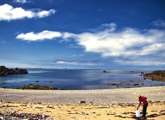 A person picking up rubbish on a sandy beach in a bay surrounded with rocks. the sea & sky are blue and there as a few clouds.